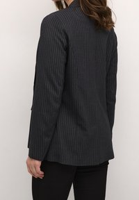 Op maat gemaakte zwarte blazer met een streepdessin van textuurstof. Beschikt over een enkele opening, opstaande revers en een getailleerde pasvorm.