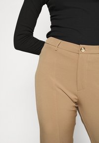 Calças beige de corte ajustado com uma textura suave, apresentando um fecho de botão único e bolsos frontais. Top preto de manga longa canelado parcialmente visível.