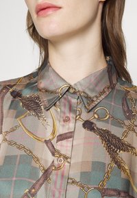 Skräddarskjorta i rutig design med guldkedjor, fjädrar och olika hårdvarudetaljer. Färgerna inkluderar beige, teal och brunt.