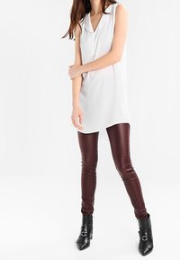 Túnica blanca sin mangas con cuello, combinada con leggins de cuero sintético burdeos ajustados y botas negras de tobillo con hebillas.