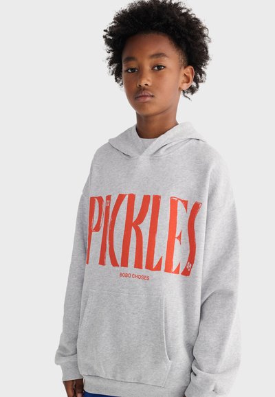 Niño con cabello rizado que lleva una sudadera con capucha gris con un gran texto rojo "PICKLES" y un bolsillo delantero, de pie contra un fondo claro y liso.