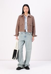 Bruine cropped jas met grote voorzakken, gedragen over een lichtblauwe blouse. Lichtblauwe flare jeans en een zwart-wit handtas maken de look compleet.