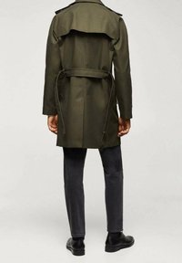 Olivgrüner Trenchcoat mit einem Gürtel, Schulterkläppchen und einem Rückenschlitz, kombiniert mit dunkelgrauen Jeans und schwarzen Schuhen.