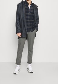 Giacca impermeabile navy, camicia a quadri con tonalità blu e grigie, pantaloni tuta grigi a gamba stretta e scarpe da ginnastica bianche.