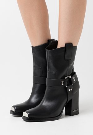 Bottes mi-mollet en cuir noir avec bouts en métal, sangles cloutées, talons carrés, et logo "GUESS" sur le talon, portées sur jambes nues.