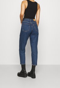 Vaqueros de denim azul con cintura alta, piernas estrechas y dos bolsillos traseros. Combinados con botas negras hasta el tobillo, mostrados desde atrás.
