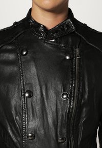 Svart läderjacka med hög krage, dubbelknäppt fram, glänsande knappar och dragkedja i sidan; slät textur med subtil lyster.