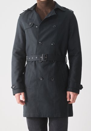 Mørk marineblå dobbeltradet trenchcoat med bælte båret over en brun rullekrave, med detaljer af knapper foran og på ærmet.