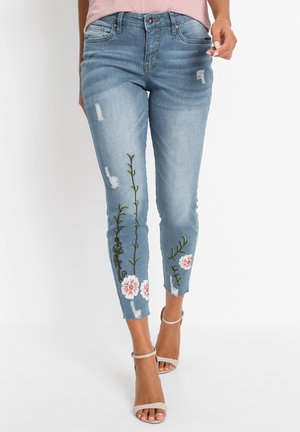 Donna che indossa jeans azzurri slavati con ricamo floreale sulle gambe inferiori e tacchi beige con punta aperta e cinturino alla caviglia.