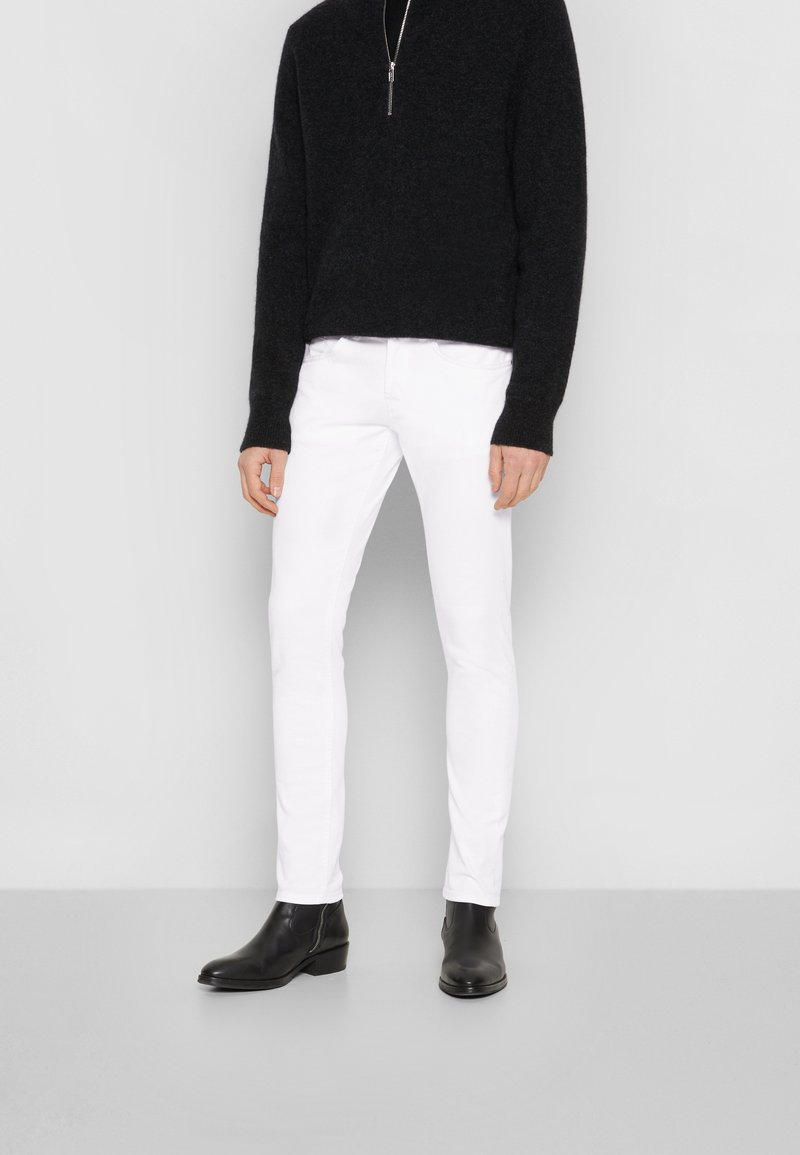 Suéter negro con cuello de cremallera, combinado con jeans ajustados blancos y botines negros. Las texturas suaves y los colores contrastantes crean un aspecto moderno.