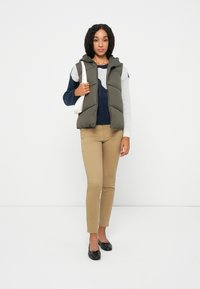 Grüne Steppweste mit Kapuze, navy und grau farblich abgesetzter Pullover, hellbeige Slim-Fit-Hose und schwarze Ballerinas, die eine weiße Handtasche hält.