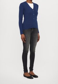 Blå ribbad cardigan med V-ringning och knappstängning, parat med mörka slim-fit jeans och svarta lägenheter, presenterad mot en enfärgad bakgrund.