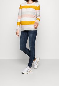 Gestreifter Strickpullover in mehreren Farben mit Gelb, Pink, Grau und Weiß, kombiniert mit dunkelblauen engen Jeans und weißen Sportschuhen.