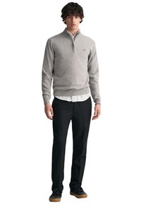 Grau zipfeliger Pullover aus weichem Stoff, kombiniert mit einem weißen Hemd und schwarzen Hosen. Das Ensemble wird mit schwarzen Sneakers abgerundet.