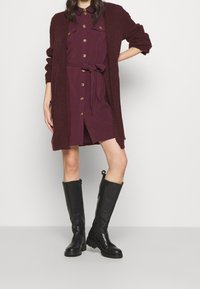 Burgundowa sukienka z guzikami i wiązaniem w talii, połączona z teksturowanym burgundowym kardiganem. Czarne skórzane kozaki za kolano z masywną podeszwą.