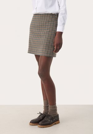 Mini-jupe à carreaux bruns en tissu tissé, avec une coupe ajustée. Associée à des chaussures foncées et des chaussettes grises texturées.
