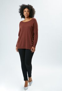 Brauner Pullover mit weitem Halsausschnitt, langen Ärmeln und seitlichen Knöpfen; kombiniert mit schwarzen Leggings und silbernen Glitzer-High Heels. Glatte Textur.