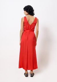 Red satin maxi dress with a V-back and a fitted waist. The dress has a smooth texture and flows to the floor, paired with leopard-print shoes.