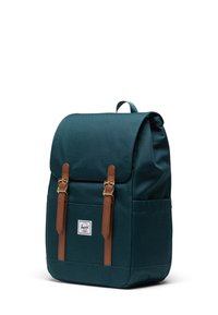 Sac à dos en toile teal avec fermeture à rabat, sangles en cuir marron, quincaillerie dorée et une poche avant avec un patch logo.
