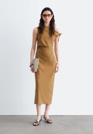 Mujer con un vestido midi marrón sin mangas con un hombro atado, sosteniendo un bolso de mano beige, llevando gafas de sol y sandalias negras con tiras.
