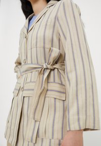 Beige gestreifter Blazer mit blauen und orangefarbenen Linien, ausgestattet mit einem gebundenen Gürtel, zwei Fronttaschen, kurzen Ärmeln und einem knopfbaren Kragen.