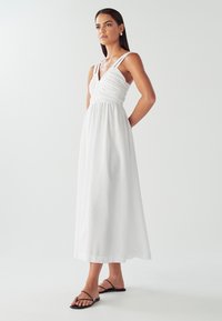 Hvid maxikjole med en figursyet overdel med rynkedetaljer, tynde skulderstropper og flydende stof. Båret med sorte sandaler med stropper.