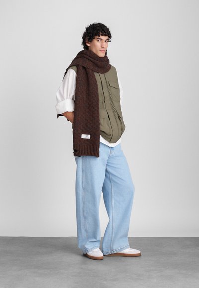 Colete cáqui com vários bolsos, lenço castanho com padrão, jeans azuis largos e tênis brancos com solas castanhas claras. Fundo simples.