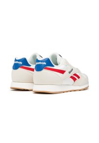 Zapatillas blancas con una textura suave, con un acento en el talón azul, rayas rojas en los laterales y una suela de goma beige. Marca en el talón.