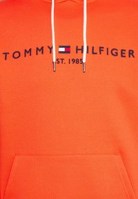 Sudadera naranja con bolsillo canguro, capucha con cordón y texto bordado "TOMMY HILFIGER EST. 1985" con detalle del logo.