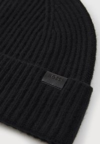 Black ribbed knit beanie made of wool, featuring a black leather label with embossed text on the folded edge. Soft texture and snug fit.