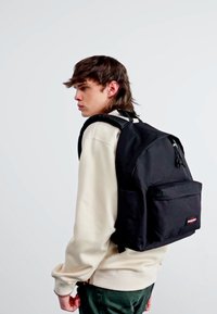 Černý bavlněný batoh s velkou hlavní kapsou, přední kapsou a nastavovacími popruhy, nošený přes svetřík světlé barvy.