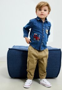 Camicia di denim con ricamo di trattore, abbinata a pantaloni cargo beige. Le calzature sono bianche con strap in velcro. Sullo sfondo si vede un pouf blu.