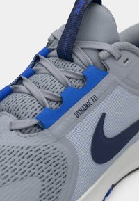 Grå idrottssko med ett meshinspirerat övre, blå accenter och en märkning för "Dynamic Fit". Har en slät yta och en framträdande marinblå Nike-logotyp.