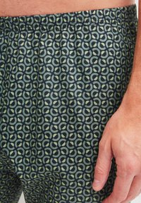 Geprinte boxershorts van stof met een donkergroene achtergrond en lichtgroene cirkelpatronen. De tailleband is elastisch en gerimpeld.
