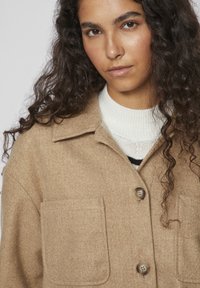 Beige Wollmischjacke mit Kragen, zwei vorderen Taschen und vorderer Knopfverschluss, darüber liegt ein weiß-schwarz gestreiftes Sweatshirt.