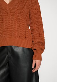 Sweter w kolorze rdzy, wykonany na drutach, z dekoltem w serek i teksturowanym wzorem w zygzaki, w połączeniu z czarnymi skórzanymi spodniami. Posiada ściągacze przy mankietach i u dołu.