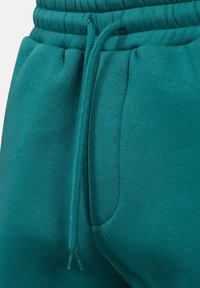 Pantalones de chándal en color verde azulado de algodón con cintura elástica, detalle de cordón y bolsillo lateral, que cuentan con una textura suave y un acabado acanalado.