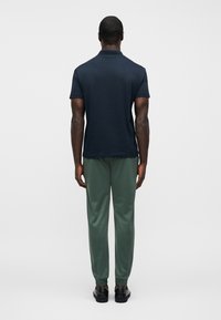 Μπλε ναυτικό μπλουζάκι πόλο, κοντά μανίκια, μείγμα βαμβακιού, συνδυασμένο με πράσινο παντελόνι με στενή γραμμή, ελαστικές μανσέτες και μαύρα παπούτσια. Το μοντέλο φαίνεται από πίσω.