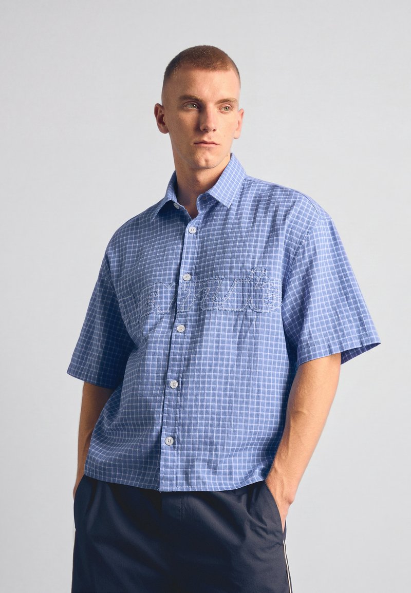 Jeune homme aux cheveux courts portant une chemise bleue à carreaux à manches courtes boutonnée et un pantalon noir, les mains dans les poches, regardant de côté.