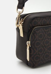 Tmavohnědá kožená kabelka s embosovaným vzorem loga CK, zlat�ými zipy a kováním. Kompaktní design s nastavitelným popruhem.