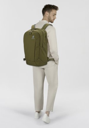 Zaino verde oliva con spallacci imbottiti, molteplici scomparti, tessuto texturizzato e dettagli con logo. Indossato da un modello in abbigliamento chiaro.