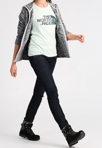 Persona che cammina indossando una giacca grigia con zip, una maglietta chiara con il logo "The North Face", jeans scuri e scarponi da trekking neri.