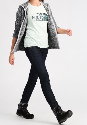 Persona che cammina indossando una giacca grigia con zip, una maglietta chiara con il logo "The North Face", jeans scuri e scarponi da trekking neri.