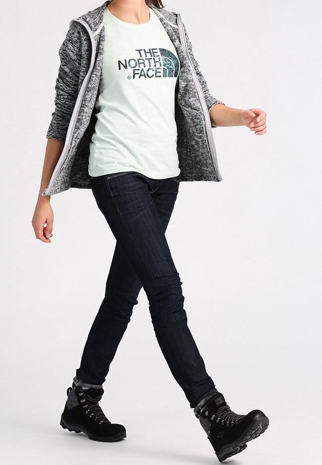 Persona che cammina indossando una giacca grigia con zip, una maglietta chiara con il logo "The North Face", jeans scuri e scarponi da trekking neri.