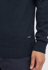 Pull en maille bleu marine avec des poignets et un ourlet côtelés, arborant une petite étiquette de marque sur le côté inférieur. Texture douce, design à coupe régulière.