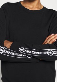 Personne portant un haut à manches longues noir avec un ruban logo "Michael Kors" courant le long des manches, les bras croisés sur la poitrine.