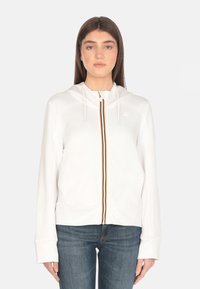 Sweat à capuche zippé blanc avec fermeture éclair à l'avant, orné d'une élégante bande colorée et d'un cordon de serrage à la capuche. Tissu doux avec des manches longues.