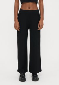 Črne široke hlače z rebrasto tkanino in visokim pasom imajo teksturiran material. Kombinirane so s črnimi čevlji z debelim podplatom za kontrastni videz.