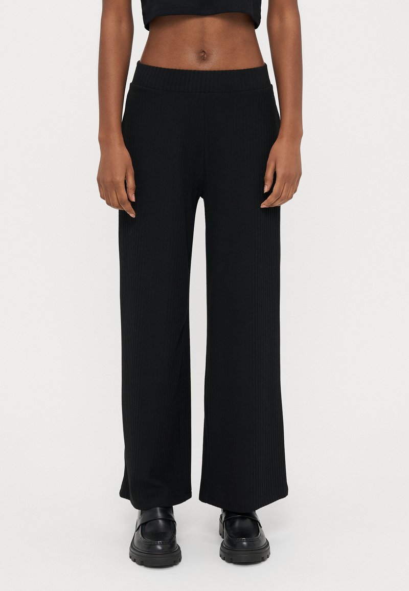 Črne široke hlače z rebrasto tkanino in visokim pasom imajo teksturiran material. Kombinirane so s črnimi čevlji z debelim podplatom za kontrastni videz.