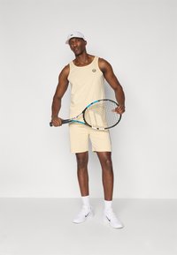 Beige linne och shorts, vit keps, hållande ett tennisracket med blå detaljer. Med en rund logotyp på toppen. Sneaker på fötterna.